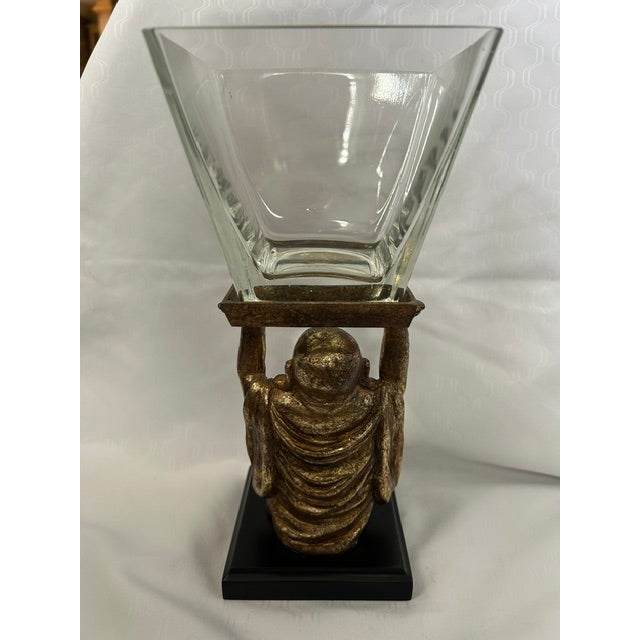 Adorable laughing Buddha holding up a glass vessel resting on a black platform. Based on a 10th-century Chinese monk, the...