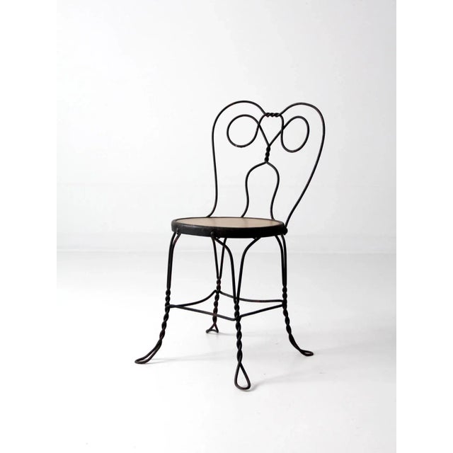 A vintage ice cream parlor chair in black metal, featuring a wide back with the classic “spectacle” design and a wood...