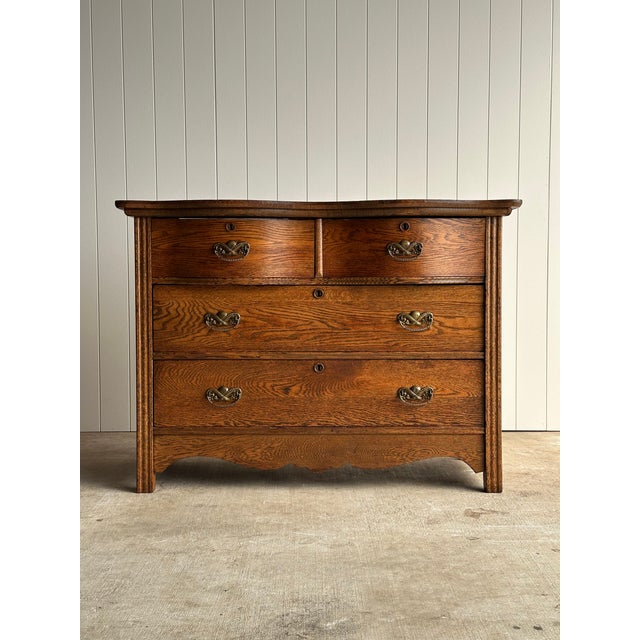 This late 19th century oak chest of drawers features a sculpted serpentine facade, shaped top, and pronounced cathedral...