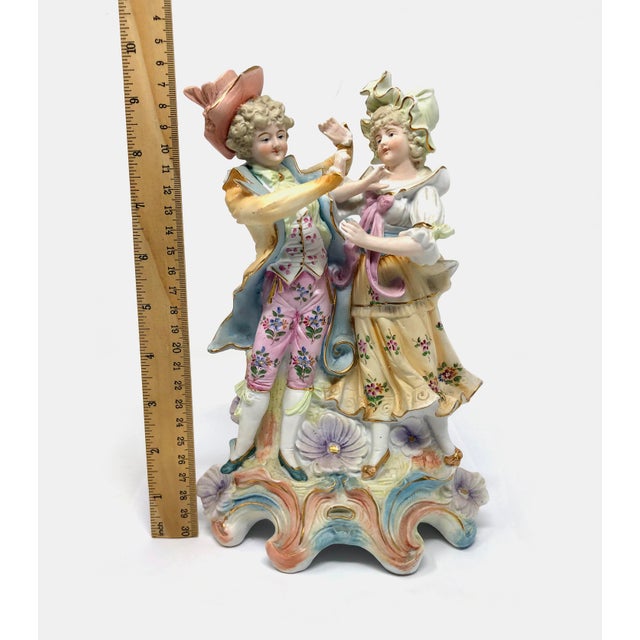 This elegant 9.5-inch tall ceramic bisque figurine depicts a courting couple in ornate, 18th-century style attire. The...