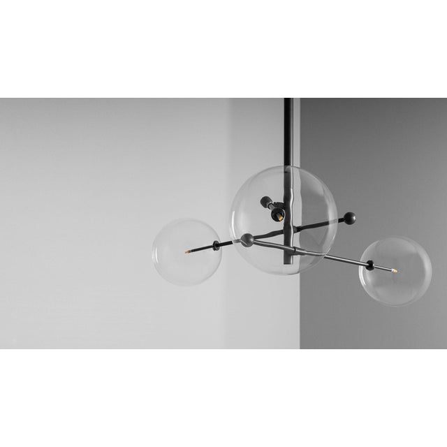 A trio of mouth-blown glass globes orbit a central axis, poised on adjustable cantilevered brass arms. The modular...