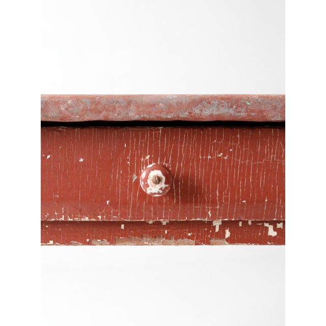 An antique farmhouse work table featuring a painted galvanized metal top with a richly layered red surface, full of depth...