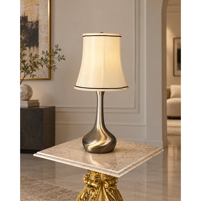 A stylish contemporary table lamp featuring a sculptural silver-tone base paired with a cream tapered shade accented by...