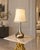 A stylish contemporary table lamp featuring a sculptural silver-tone base paired with a cream tapered shade accented by...