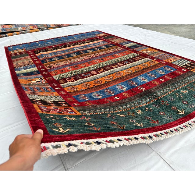 This hand-knotted Khorjin rug features a vivid patchwork of horizontal bands in rust, blue, gold, and green, each filled...