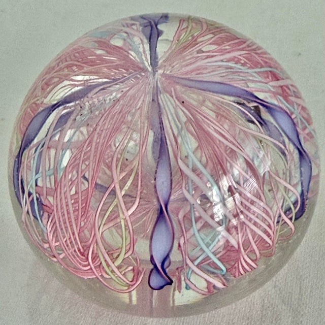 Hand blown clear glass paperweight in a round dome shape. Inside, twisted pink latticino threads swirl around lavender...