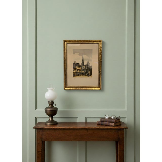 This is a hand-colored framed etching, depicting a European village or city street scene with a prominent church spire. It...