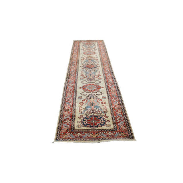 This traditional hand-knotted runner rug features an elongated ivory field anchored by a repeating, interconnected...