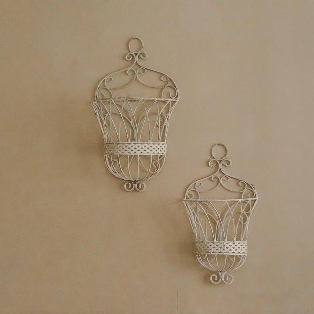 Two-piece set of decorative wall baskets made of white-painted metal with curved wire ornamentation. The design is...