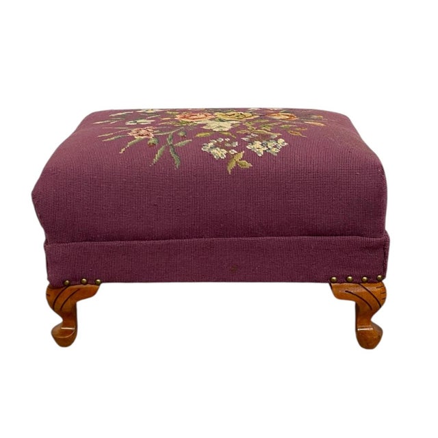 This upholstered footstool features a richly colored textile surface with a detailed floral needlepoint design set against...