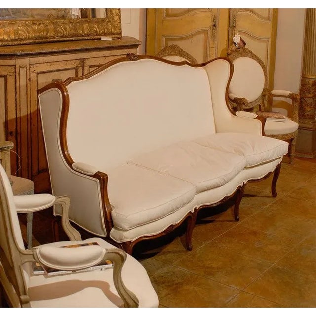 A French Louis XV style three-seat wooden winged sofa from the 19th century with carved crest, scrolled arms, cabriole...