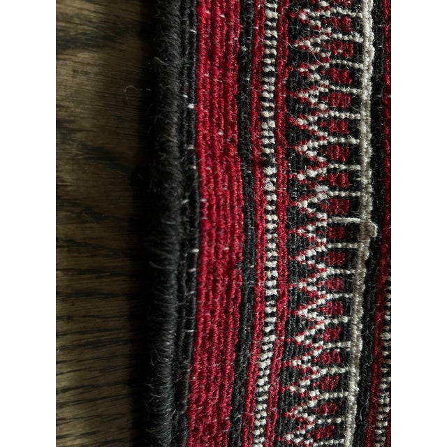 A striking handwoven rug featuring a rich and vibrant palette of deep red, burgundy, black and ivory. Its intricate...
