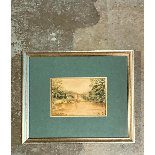 This vintage framed landscape presents a tranquil watercolor scene of a stone bridge spanning a gentle river, rendered in...