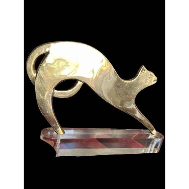 A brass cat sculpture on a clear acrylic base stylized with a smooth, elongated body and a tail curled under. The cat is...