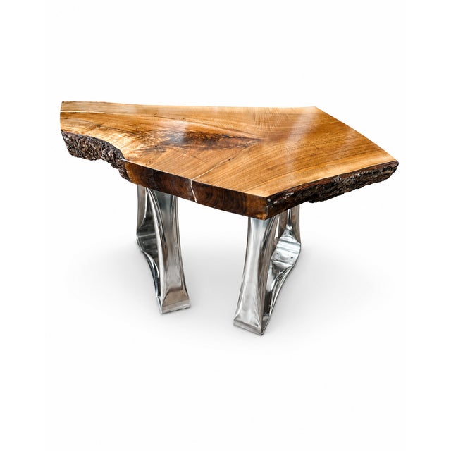 A striking contemporary live edge table featuring a richly figured natural wood slab supported by sculptural polished...