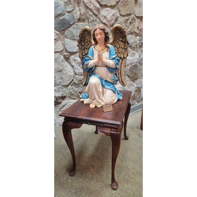 This kneeling angel joins both hands in prayer, framed by large gold-toned feathered wings. The white robe, blue mantle...