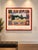 Framed art print of Kate Barnes’ “American Odyssey”: a pastoral farmhouse landscape inside a patchwork border of quilt-...