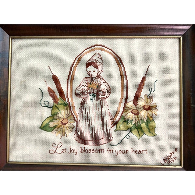 This piece is a framed cross-stitch embroidery sampler, dated 1986. The artwork features a charming, folk-art-inspired...