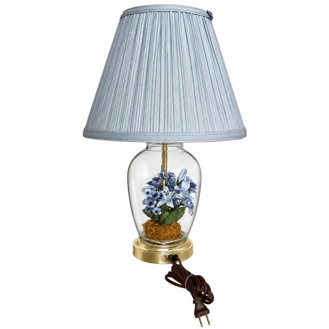 A charming floral table lamp featuring a blue botanical arrangement enclosed within a clear, vase-shaped glass body. Lily-...