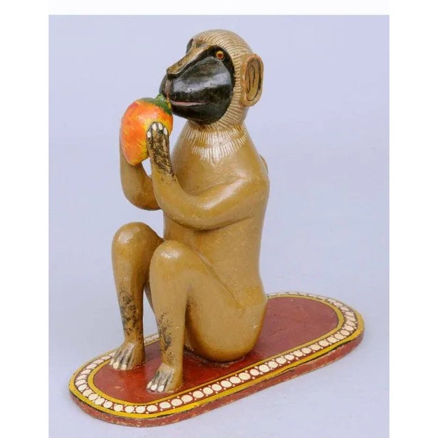 Whimsical Indian carved wood and polychromed monkey sitting on a red oval carpet with white dotted border eating a mango.