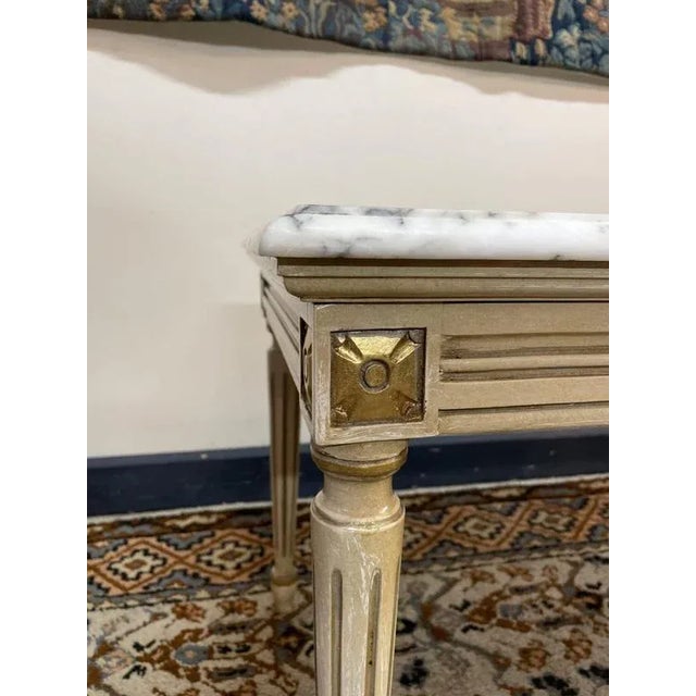 Elegant neoclassical coffee table with a veined marble top, resting on tapering legs. The frame is finished in a soft...