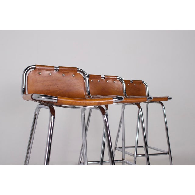 Selection in the style of charlotte perriand (1903-1999). Set of three low-back bar stools, cognac leather riveted to a...