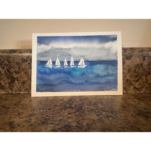 Five sailboats glide peacefully across deep blue waters beneath a moody sky in this serene watercolor seascape. The...