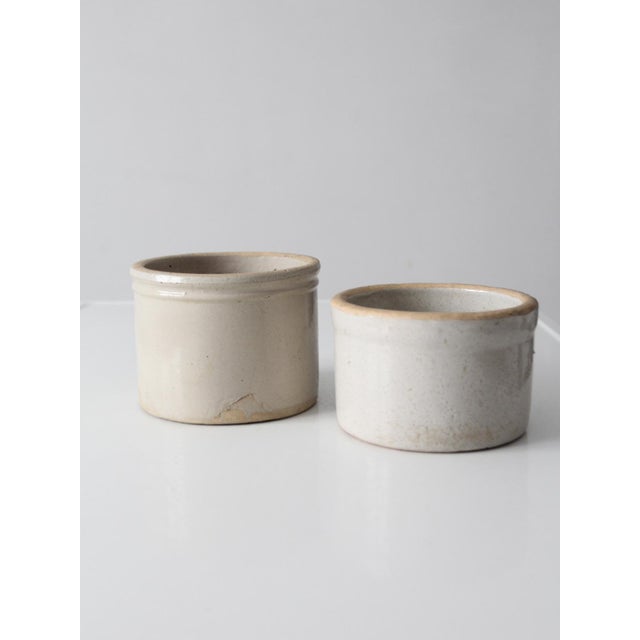 Pair of antique white stoneware crocks with creamy glaze, rounded rims, and classic cylindrical forms. Subtle glaze...