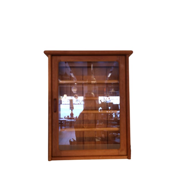 A neat wall-mounted cabinet in teak with lighting and a door in teak and glass. The cabinet has soft and elegant shapes...
