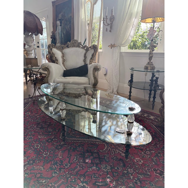 Striking vintage Hollywood Regency two-tier oval coffee table featuring sculptural glass supports, gilt metal accents, and...
