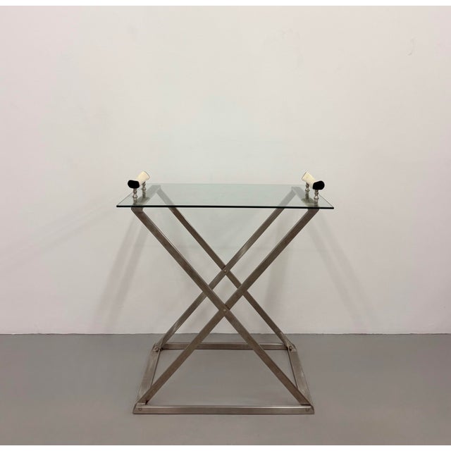 A sleek vintage serving table featuring a clear tempered glass top supported by a brushed stainless steel X-form base....