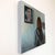 This oil painting depicts a figure with their back turned, gazing contemplatively at a portrait on the wall. The muted...