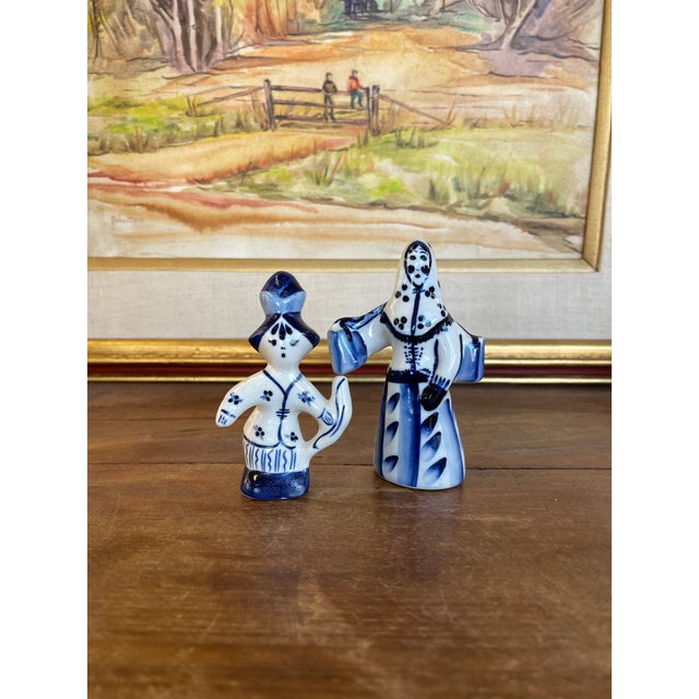 A pair of vintage blue and white porcelain figurines depicting stylized folk figures in traditional dress. Each figure is...