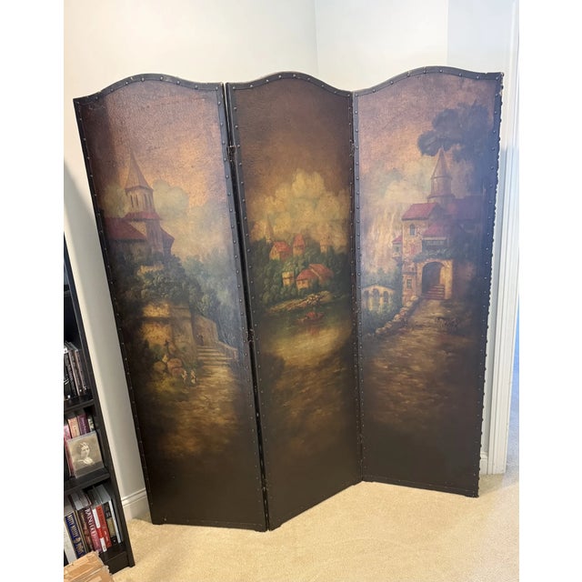 Antique painted leather screen depicting a medieval village scene and enhanced with a black iron rivet trim. Front of...