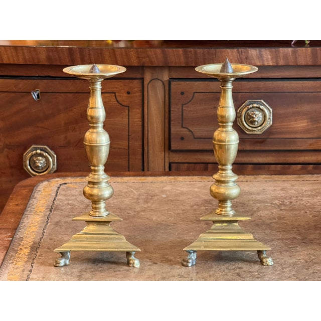 Elegant brass altar sticks featuring baluster columns rising from stepped triangular bases set on claw feet. The surfaces...