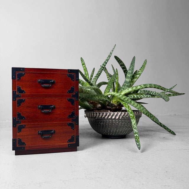 A compact Japanese storage cabinet inspired by traditional tansu design, with three drawers, warm reddish-brown wood and...