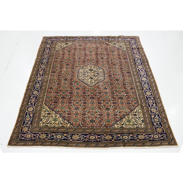 This Persian hand-knotted wool rug has a terracotta color field and accents in green and blue throughout the piece. This...