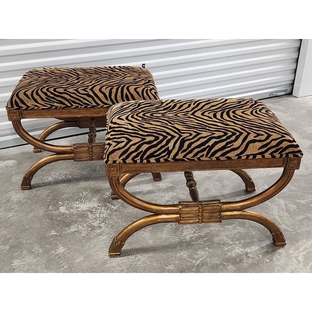 Pair of decorative carved wood uphostered curule or X-benches in a Neoclassical style. The benches feature an antique gold...