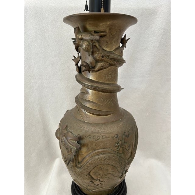An antique bronze table lamp wrapped with raised dragons in relief circling the lamp. The Asian style lamp sits on a round...