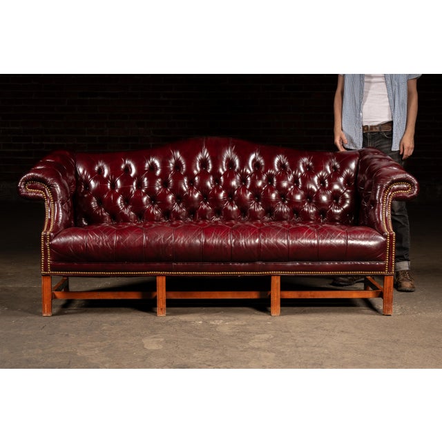 Hand-tufted oxblood leather camelback sofa with rolled arms, brass nailhead trim, and a solid mahogany base frame. The...