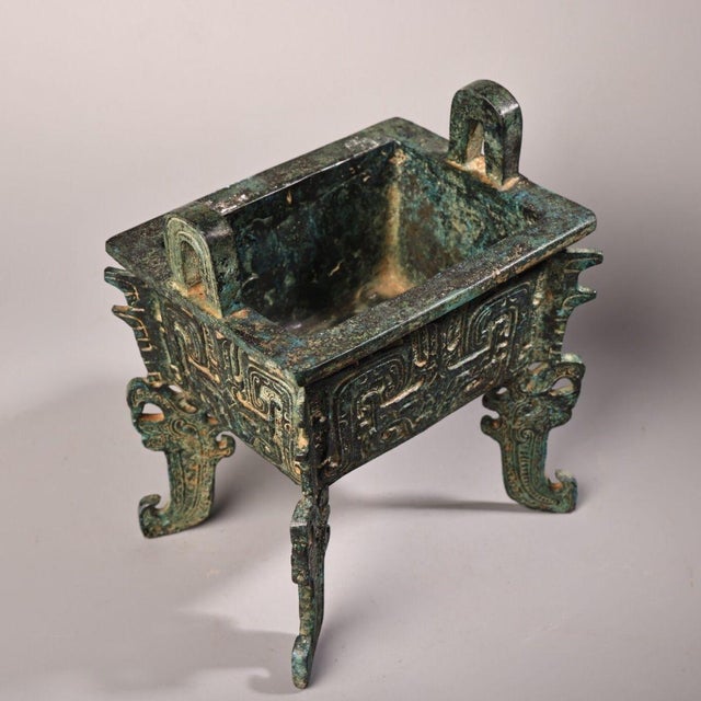 This is a cast bronze rectangular ritual vessel, featuring a sturdy, box-like body supported by four distinctive legs and...
