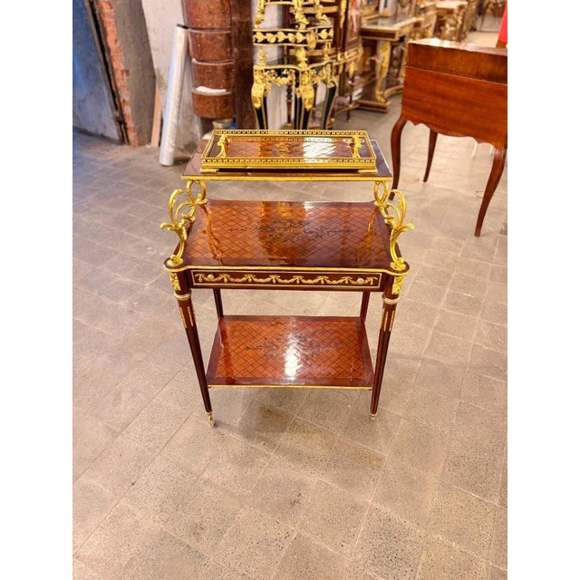 A striking French Louis XVI-style two-tier serving table featuring rich mahogany tones and beautifully detailed gilt...