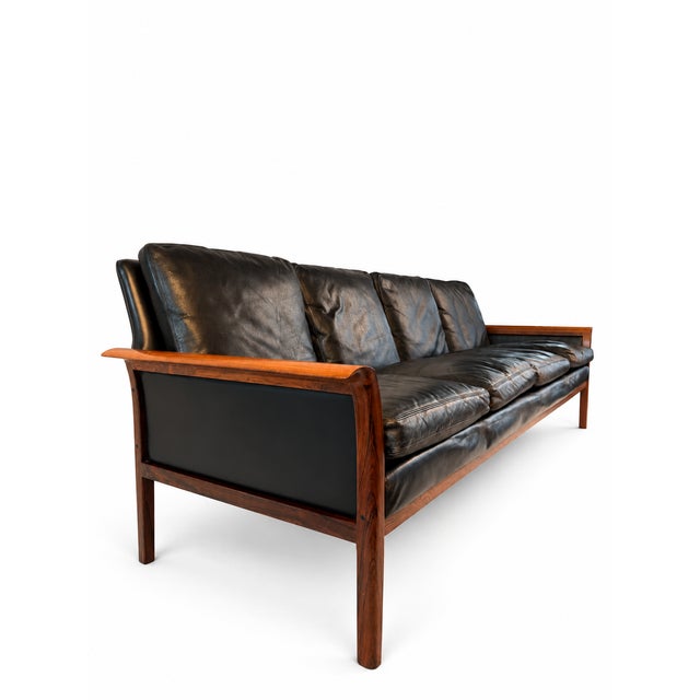 A long, low Hans Olsen rosewood and black leather sofa. The exposed wooden frame creates a warm outline around four loose...