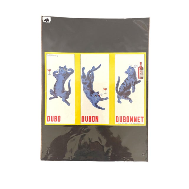 This French advertising print features the iconic Dubonnet aperitif design with a whimsical blue cat in three playful...
