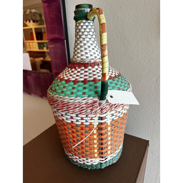 A richly colored vintage demijohn bottle wrapped in handwoven orange, green, and white vinyl, bringing texture,...