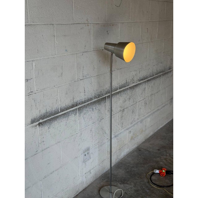 This sleek metal floor lamp features a slender stem and a flexible gooseneck arm supporting a directional shade. It...