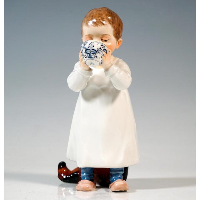 Boy in a nightgown standing in front of a damaged wooden horse, holding an onion pattern cup with both hands and drinking...