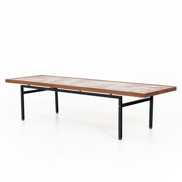 A modernist cocktail table with an inset decorative top surrounded by wooden frame. Resting a black metal base.