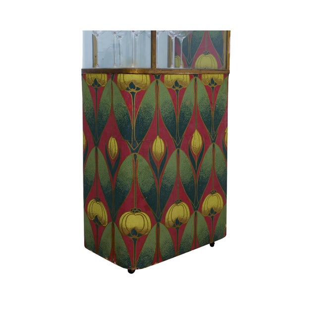 A striking Art Deco vitrine with a brass frame, curved glass panels, and bold patterned sides in red, green, and gold. The...