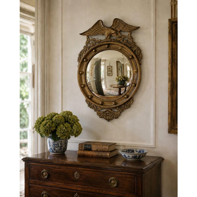 A Federal-style giltwood convex mirror crowned with a carved eagle, its wings outstretched above a laurel motif—an...